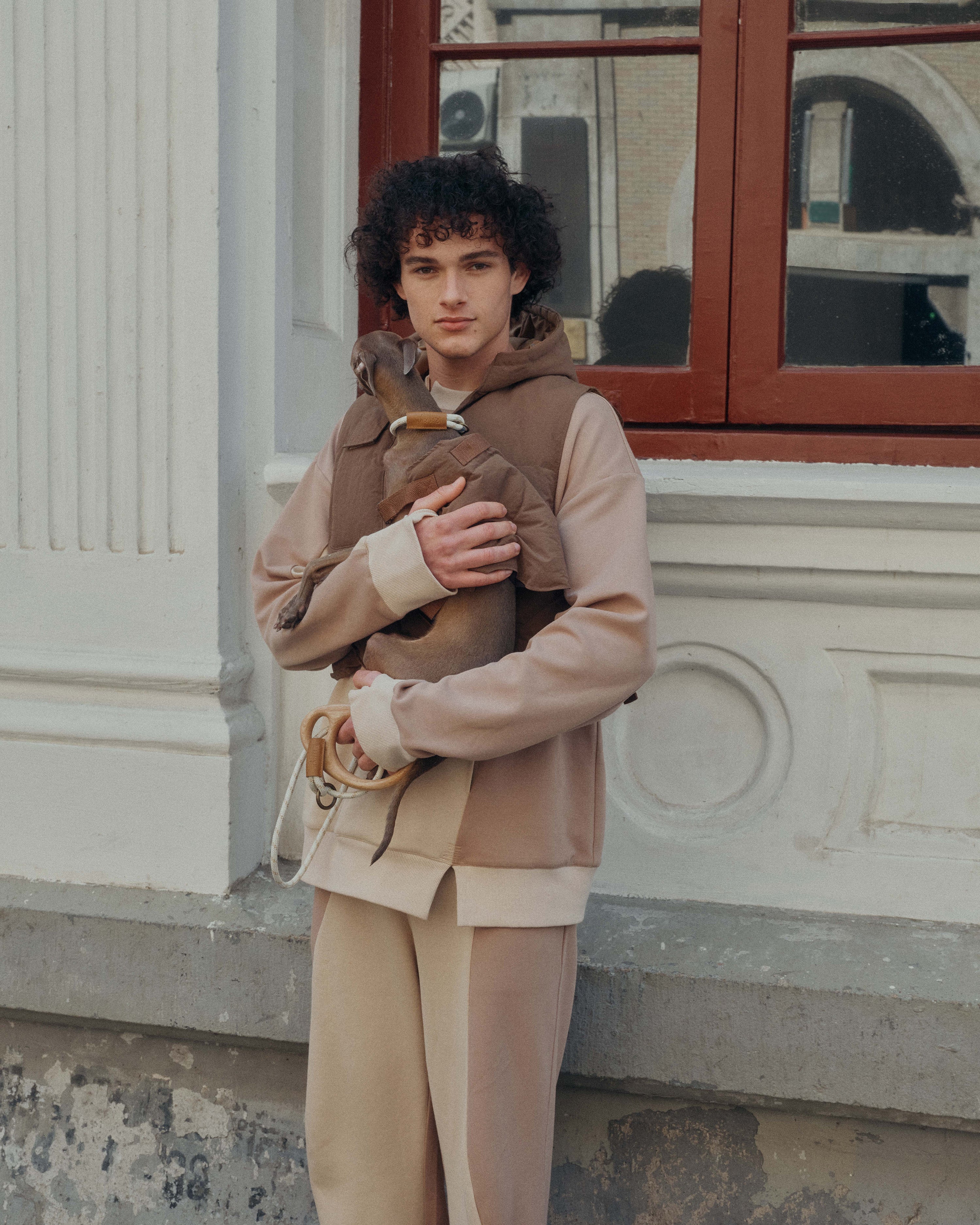 A male model holds a dog in front of windows, both of them wear patchwork sweatshirt sets.