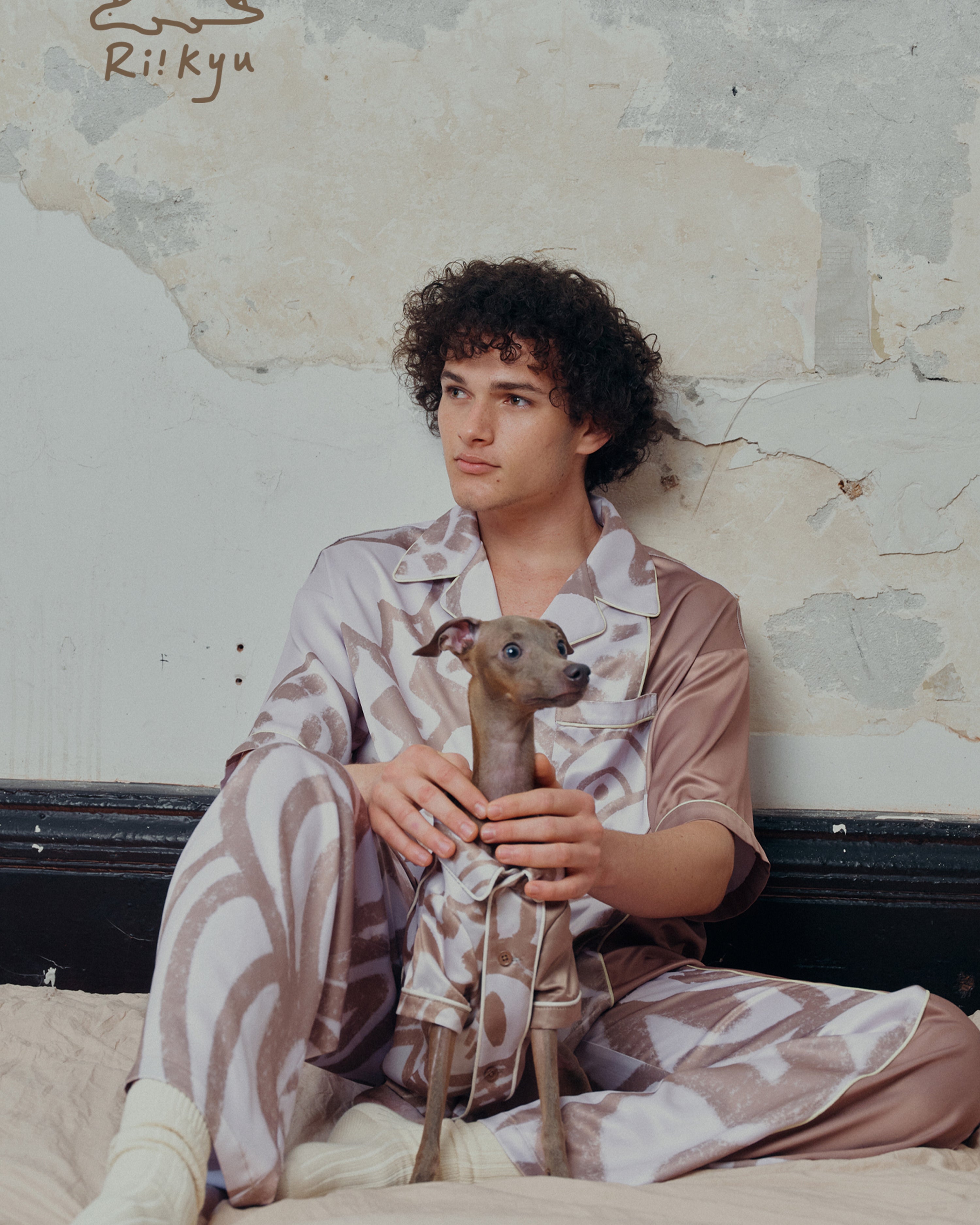 A male model sits on a bed with his dog, both the owner and the dog wear short sleeve pajama sets.