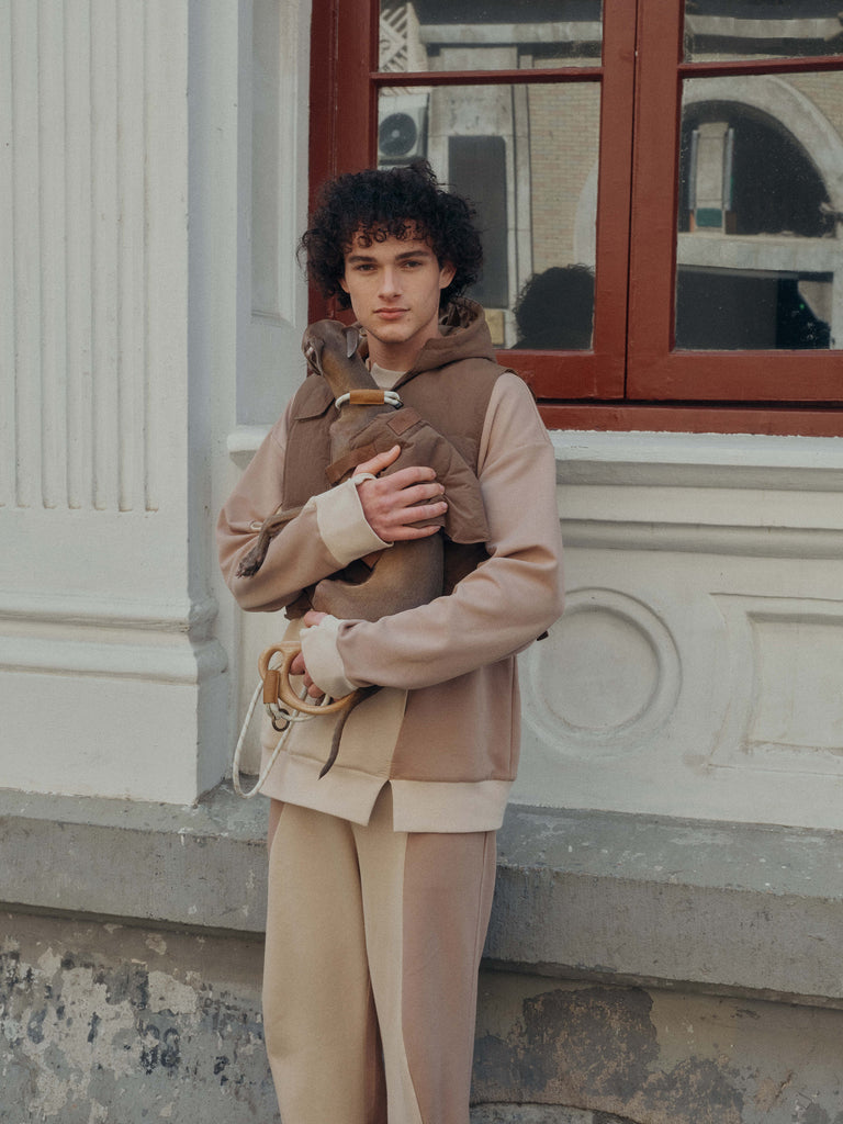 A male model holds a dog in front of windows, both of them wear patchwork sweatshirt sets.