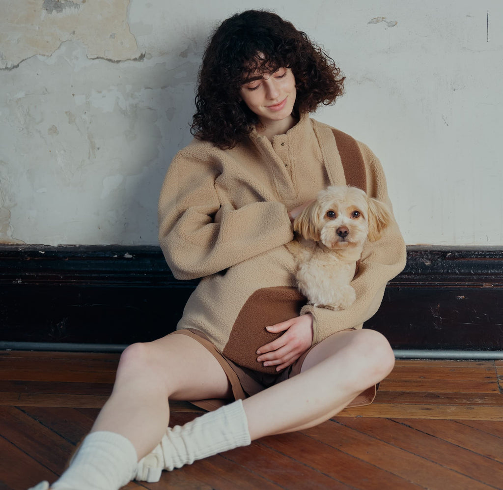 A female model sits in an oversized fleece with a dog in her  pouch.