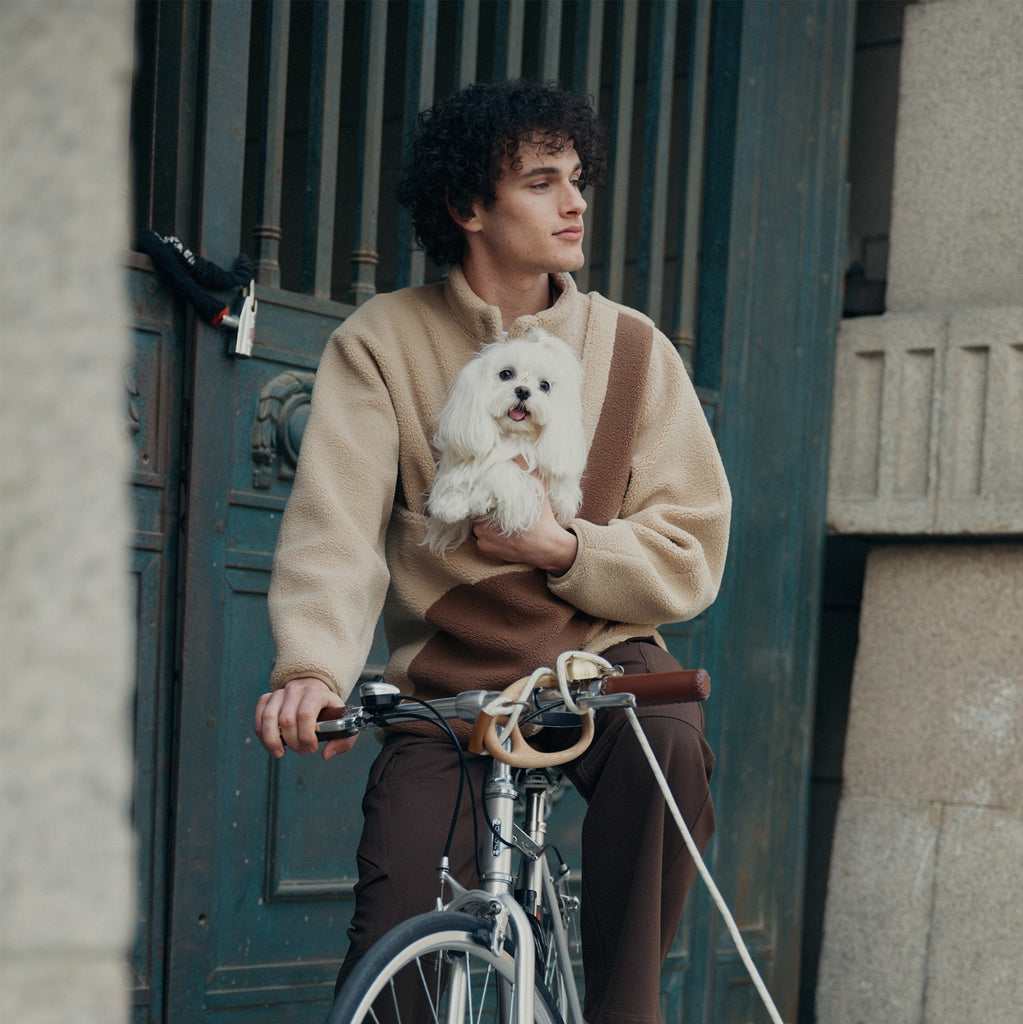 A male model is sitting in a car with a dog in his pouch. And a "Pebble" pet leash is fixed on the handlebar.